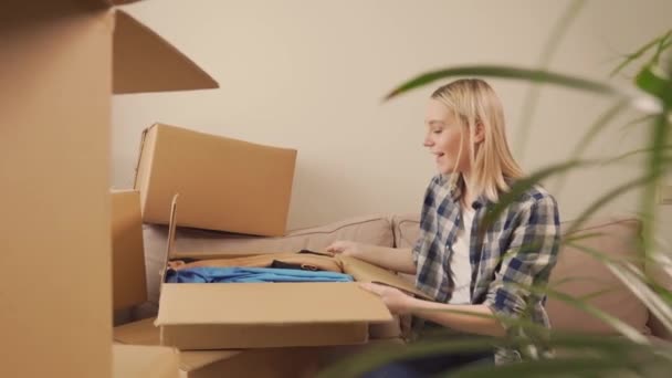 Женщина, сидящая на диване, вынимает одежду из картонной коробки после переезда — стоковое видео