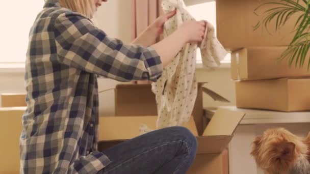 Женщина разбирает коробки с одеждой после переезда. Рядом маленькая собачка. — стоковое видео