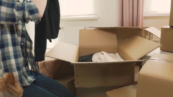 Una mujer con una camisa a cuadros pone ropa en cajas de cartón, mudándose a una casa nueva. — Vídeo de stock