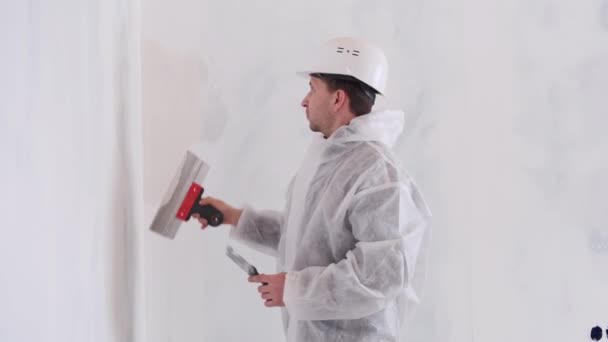 A man in a protective suit levels the walls with a spatula and plaster. — Vídeo de Stock