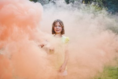 Kısa saçlı bir kız renkli dumanın içinde duruyor ve kameraya bakıyor..