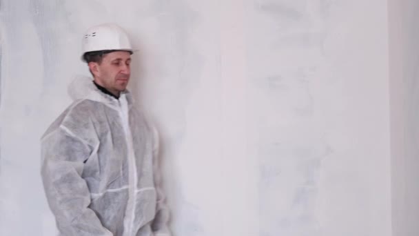 Un homme en combinaison de protection et un casque tient un rouleau pour peindre les murs. — Video