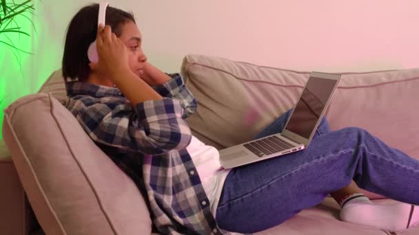 Mladá žena ležící na gauči s laptopem a poslouchající hudbu. — Stock video