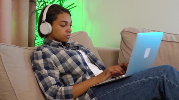 Μια γυναίκα με ένα casual πουκάμισο με ακουστικά, κρατώντας και χρησιμοποιώντας ένα ασημί laptop — Αρχείο Βίντεο