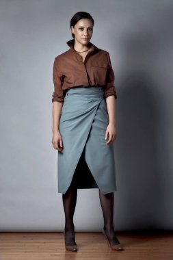 Stüdyoda kahverengi bir bluz ve uzun gri bir etekle poz veriyor..