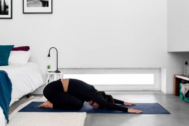 Genç, sıska bir kadın evdeki minimalist yatak odasında uzun çocuk pozisyonunda yoga yapıyor.