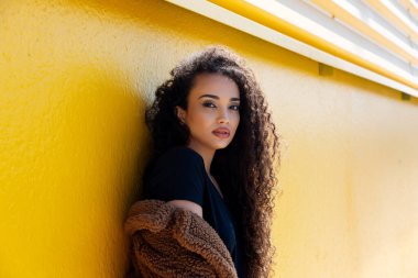 Uzun kıvırcık saçlı ve sarı duvarlı kahverengi paltolu siyah kadın.