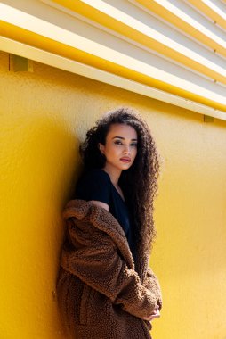 Uzun kıvırcık saçlı ve sarı duvarlı kahverengi paltolu siyah kadın.