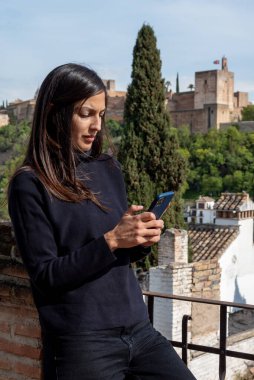 Hispanic female tourist messaging on mobile phone while standing on terrace with views to Alhambra fortress in Granada, Spain