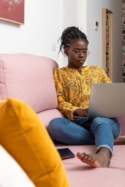 Odaklanmış Afrikalı Amerikalı kadın serbest yazar evdeki kanepede oturuyor ve internet üzerinden proje üzerinde çalışırken daktilo yazıyor.