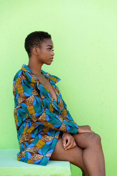 Young black female in traditional African piece of fabric looking away and sitting against pistachio background