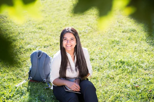 Portrait of a young Indian student, business woman. A woman with a backpack in a rucksack holds a tablet, sits on the green grass, against the sun. Summer day