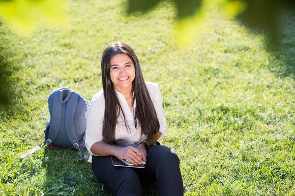 Portrait of a young Indian student, business woman. A woman with a backpack in a rucksack holds a tablet, sits on the green grass, against the sun. Summer day