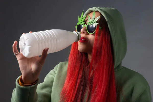 marijuana milk young hipster girl in the hood, wearing marijuana sunglasses, drinks milk from a white bottle. Cannabis and its usage. Marijuana leaf and edible marijuana products. Butter, tea, beer, energy drink, oil, chocolate, cookies and milk.