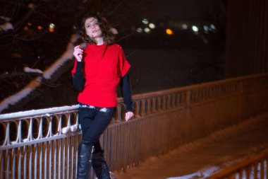 Kırmızı elbiseli, kızıl kışta poz veren model kız köprüde poz veriyor, kar yağıyor.