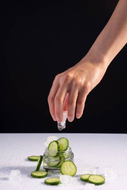 Siyah ve beyaz arka planda buz küpleri olan cam kabın içinde salatalık özü bulunan eller için ferahlatıcı doğal kozmetik ürünleri.