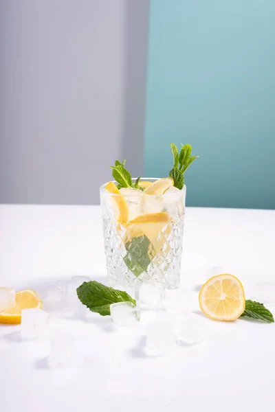 creative summer cocktail with ice in a glass glass with lemon, mint and ice cubes on a white-blue background.