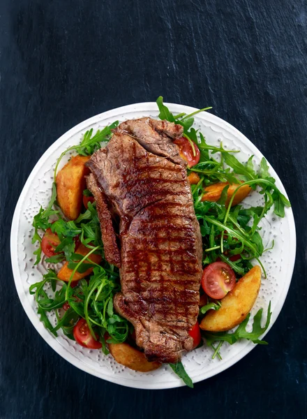 Vzácné hovězí steak s pečenými bramborami, rajčaty a rukola listy. na staré kamenné modré pozadí — Stock fotografie