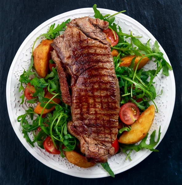 Filete de res rara con patatas asadas, tomates y hojas de cohete silvestres. sobre fondo de piedra azul viejo — Foto de Stock