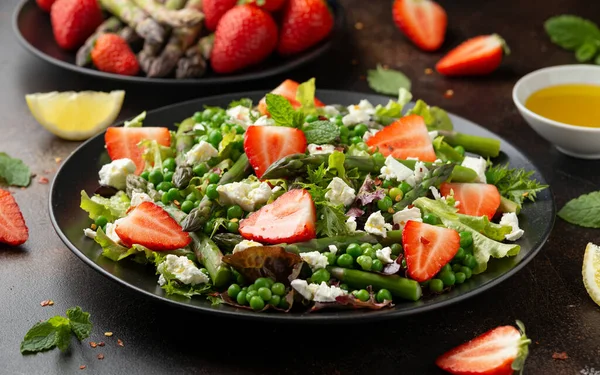 Спаржа и клубничный салат с овощами, зеленым горошком и сыром фета в черной тарелке. здоровое питание. — стоковое фото