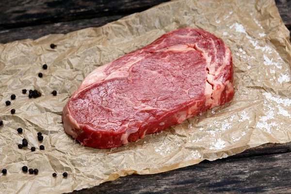 Raw rib eye steak on crumpled paper, pepper, salt. — Stok fotoğraf