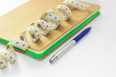 Beyaz bir yüzey üzerinde mavi kalem yanında mezura bulunan bir defter.