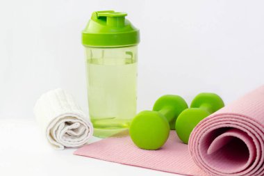 Bir şişe su, yeşil dambıl ve beyaz arka planda izole edilmiş pembe bir spor paspasın üzerine havlu. Spor malzemeleri.