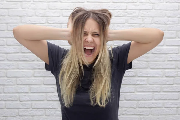 A beautiful young caucasian blonde woman holds her head, screams and covers her ears with her hands. Despair, frustration, headache, migraine, exhaustion, emotional breakdown, voices in the head, fear