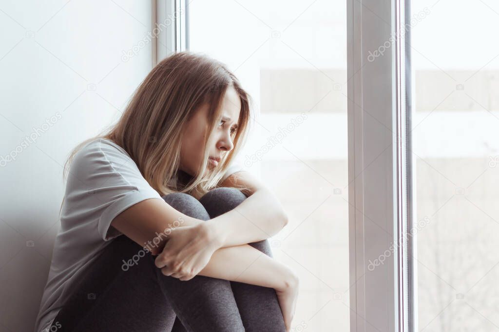 A young blonde beautiful despaired woman introvert in the white t-shirt and grey leggings is sitting on the windowsill, hugging her legs and looking out the window. Self-isolation, lock down. Solitude