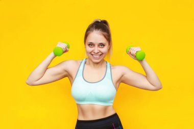 Parlak sarı arka planda izole edilmiş yeşil dambılları olan güçlü, güzel, beyaz, sarışın bir kadın. Düz karın kasları olan sportif bir kız halterle egzersiz yapar. Zindelik