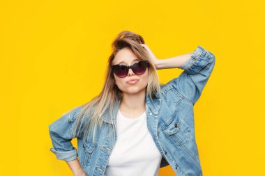 Beyaz tişörtlü, beyaz kot ceketli, siyah gözlüklü, eli parlak sarı arka planda izole edilmiş bir kafanın üzerinde duran beyaz, şık, sarışın bir kadın. Güzellik, moda