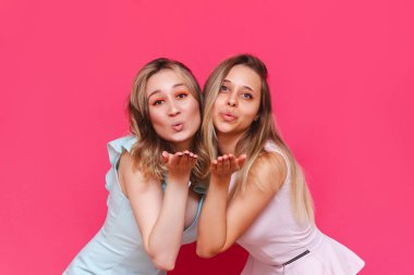 İki beyaz, şık moda elbiseli zarif kadın parlak pembe bir arka planda izole edilmiş kameraya açık avuç içleriyle hava öpücüğü gönderiyorlar. Güzel kızlar öpüşür. Arkadaşlar