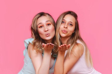 Kafkasyalı zarif elbiseli iki genç kadın açık avuçlu hava öpücüğü gönderip açık pembe arka planda izole edilmiş kameraya bakıyorlar. Güzel kız arkadaşlar öpüşür.
