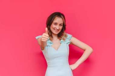 Açık mavi elbiseli, gülümseyen kahverengi saçlı güzel beyaz bir kadın. Parlak pembe bir arka planda eli izole edilmiş bir jest gibi baş parmağını gösteriyor. Olumlu insani duygu