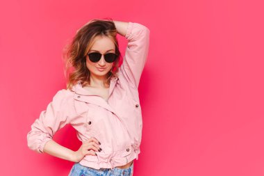 Gayri resmi bir ceket ve siyah güneş gözlüğü takmış, kendine güvenen beyaz, kahverengi saçlı genç bir kadın parlak pembe bir arka planda eli başındaymış gibi poz veriyor. Güzellik, moda. Boşluğu kopyala
