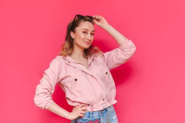 Güzel, kendine güvenen kahverengi saçlı, sıradan bir ceket ve kot şortla genç bir kadın parlak pembe bir arka planda izole edilmiş güneş gözlüğü tutarak poz veriyor. Güzellik ve moda