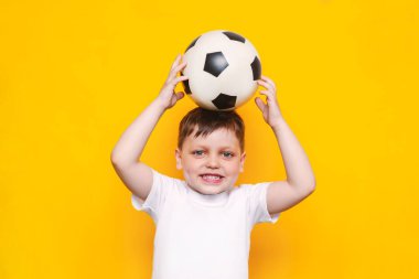 Beyaz tişörtlü küçük sevimli bir çocuk gülümsüyor ve parlak sarı arka planda izole edilmiş bir futbol topunu kafasında tutuyor. Topu beyaz tenli bir çocuk tutuyor. Çocuk sporları oyunu. Zindelik