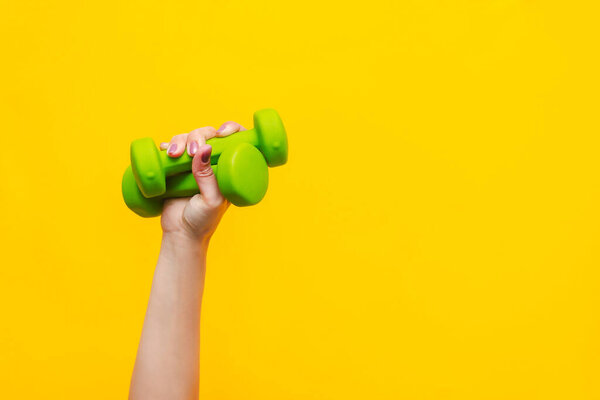 Two green dumbbells in a woman's hand isolated on yellow background. Sport equipment. Fitness concept, healthy lifestyle. Copy space for design