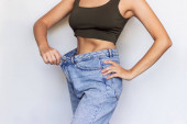 Oříznutý záběr mladé štíhlé ženy oblečené v širokých džínách a krátký top demonstruje úspěšný úbytek hmotnosti izolované na světle šedém pozadí. Dietní a wellness koncept