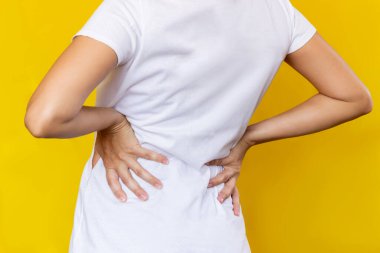 Beyaz tişörtlü, elleri sarı arka planda izole bir şekilde belini tutan genç bir kadının kırpılmış görüntüsü. Sırt ağrısı. Siyatik, osteokondroz, safra taşı hastalığı, pankreas iltihabı