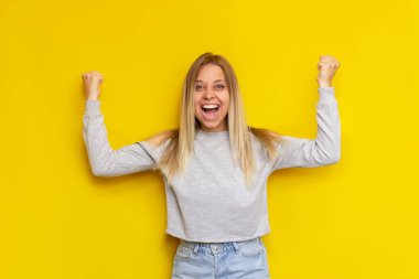 Genç, beyaz tenli, etkileyici, kolları gündelik giysiler içinde yükselen sarışın kadın, parlak sarı bir arka planda izole edilmiş haber ya da piyango kazanmasından mutlu.