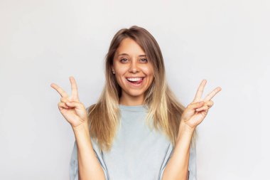 Aşırı büyük tişörtlü, gülümseyen, neşeli, neşeli, genç beyaz bir kadın dil ve beyaz bir arka planda elleri izole bir barış hareketi gösteriyor. Kız zafer işareti gösteriyor.