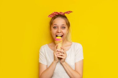 Beyaz kısa bluzlu, gülümseyen sarışın bir genç beyaz kadın parlak sarı bir arka planda izole edilmiş waffle külahında meyveli dondurma yiyor. Yaz konsepti