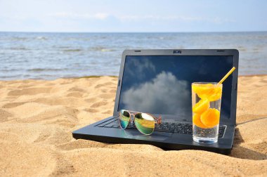 Kumsalda uzaktan kumanda, siyah bir laptop ve güneş gözlüğü..
