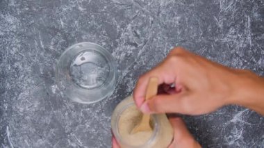 Genç bir adam şeffaf bardağa beyaz tahta kaşık kullanarak esmer şeker döküyor. Beton desenli arka planda izole edilmiş. kopyalama alanı. 4k görüntü