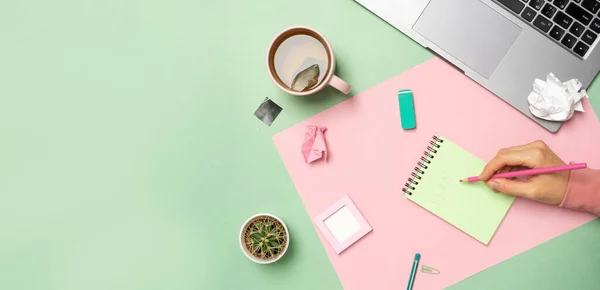 Pastel ofis masasında dizüstü bilgisayarı, not defteri ve bir kadın eli var..