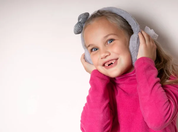 Klein schattig tandeloos meisje rommelen rond in warme bont koptelefoon op een lichte achtergrond. — Stockfoto