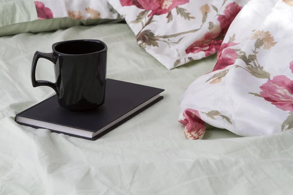 Caneca preta com um livro preto na roupa de cama florida — Fotografia de Stock