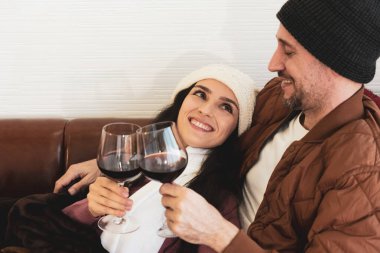 Genç beyaz bir çift, gülen yüzlü, kanepede yan yana oturan ve kışın evde kırmızı şarap içen. Mutlu aile kavramı