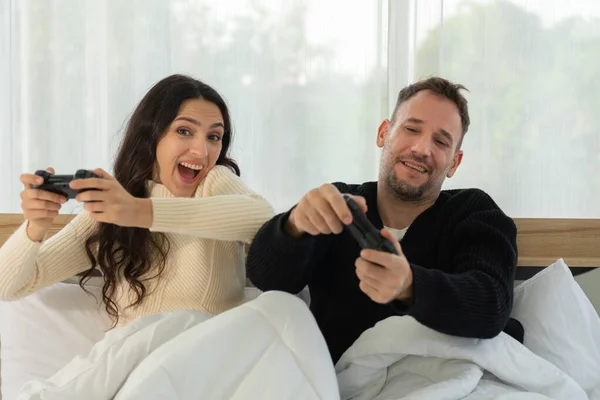 caucasian couple sitting in bed in bedroom playing virtual reality game together at home